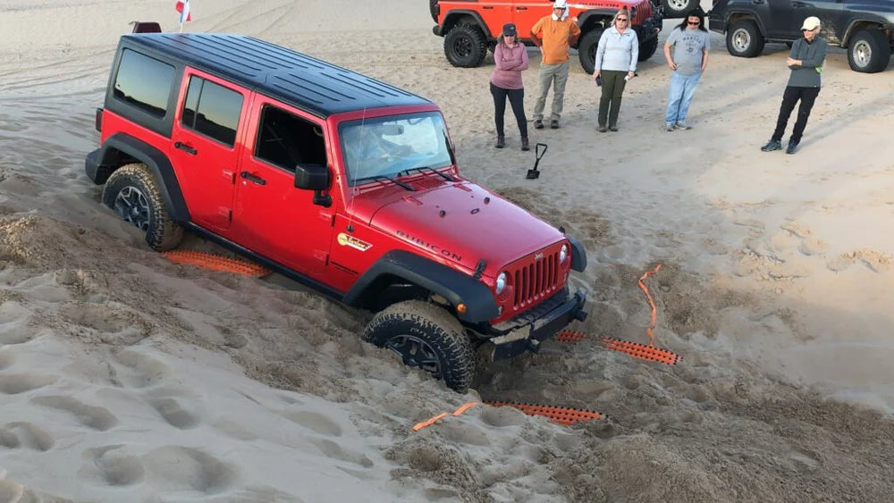 A red Jeep Wrangler stuck in deep sand, with several people standing nearby on a sandy terrain. Traction boards are positioned under each tire.