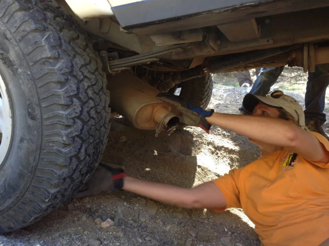 A person in an orange shirt and cap works underneath a vehicle, touching a tan exhaust pipe near a large off-road tire on a dirt ground.
