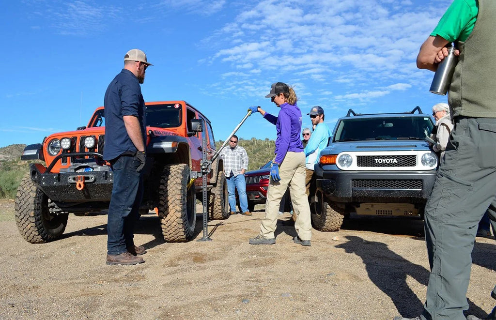 Several people standing outdoors between two off-road vehicles on a dirt lot under a blue sky. One person holds a long metal tool near the center, with an orange Jeep-style vehicle on the left and a dark SUV on the right.