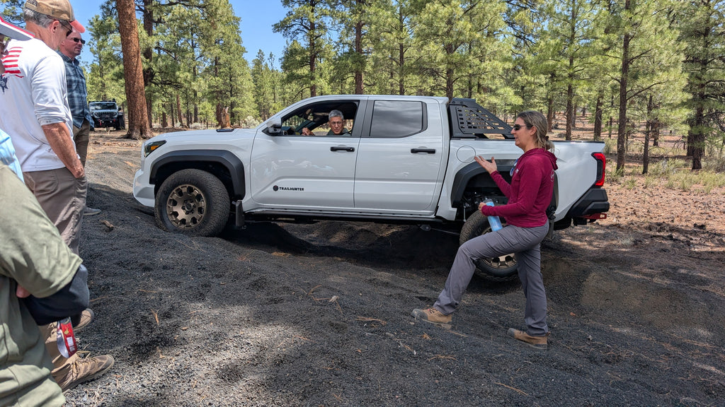 A white Toyota Tacoma pickup truck straddles a small ditch with one wheel off the ground. A small group of people are observing the articulation of the vehicle as an instructor wearing a burgundy shirt and grey pants explains what is happening.