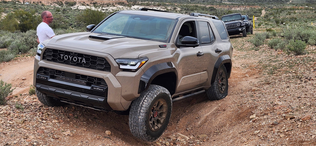 A tan Toyota 4Runner SUV parked on a dirt trail in a desert landscape, with a man standing beside it and another vehicle in the background.