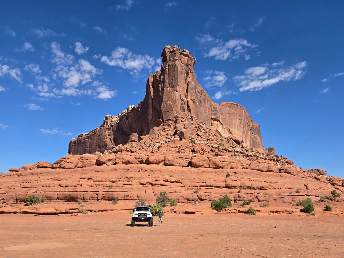 A large red rock butte rises under a bright blue sky with scattered white clouds, with a small white vehicle parked on a red dirt desert plain in the foreground.
