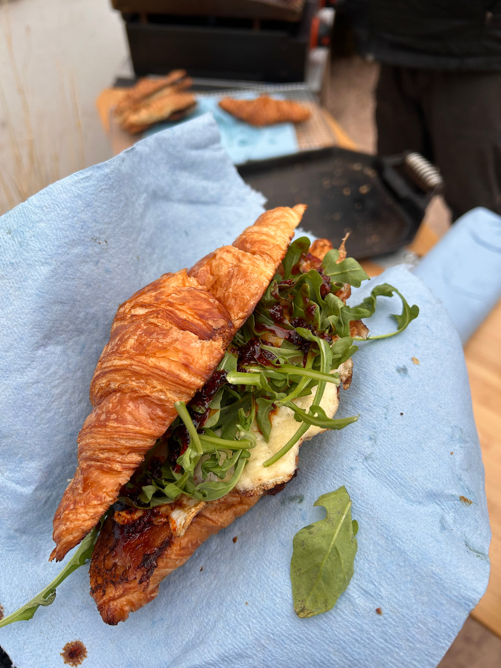 A croissant breakfast sandwich with leafy greens, melted cheese and a fried egg sits atop a blue napkin with cooking pans and other foods in the background.