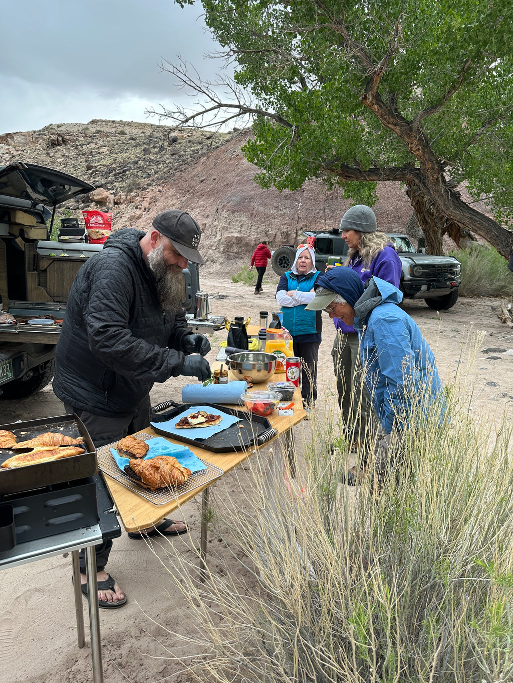 A group of people gathered outdoors at a table in a desert setting, preparing and serving food. One man in a black jacket and cap is preparing breakfast sandwiches, while others stand nearby.