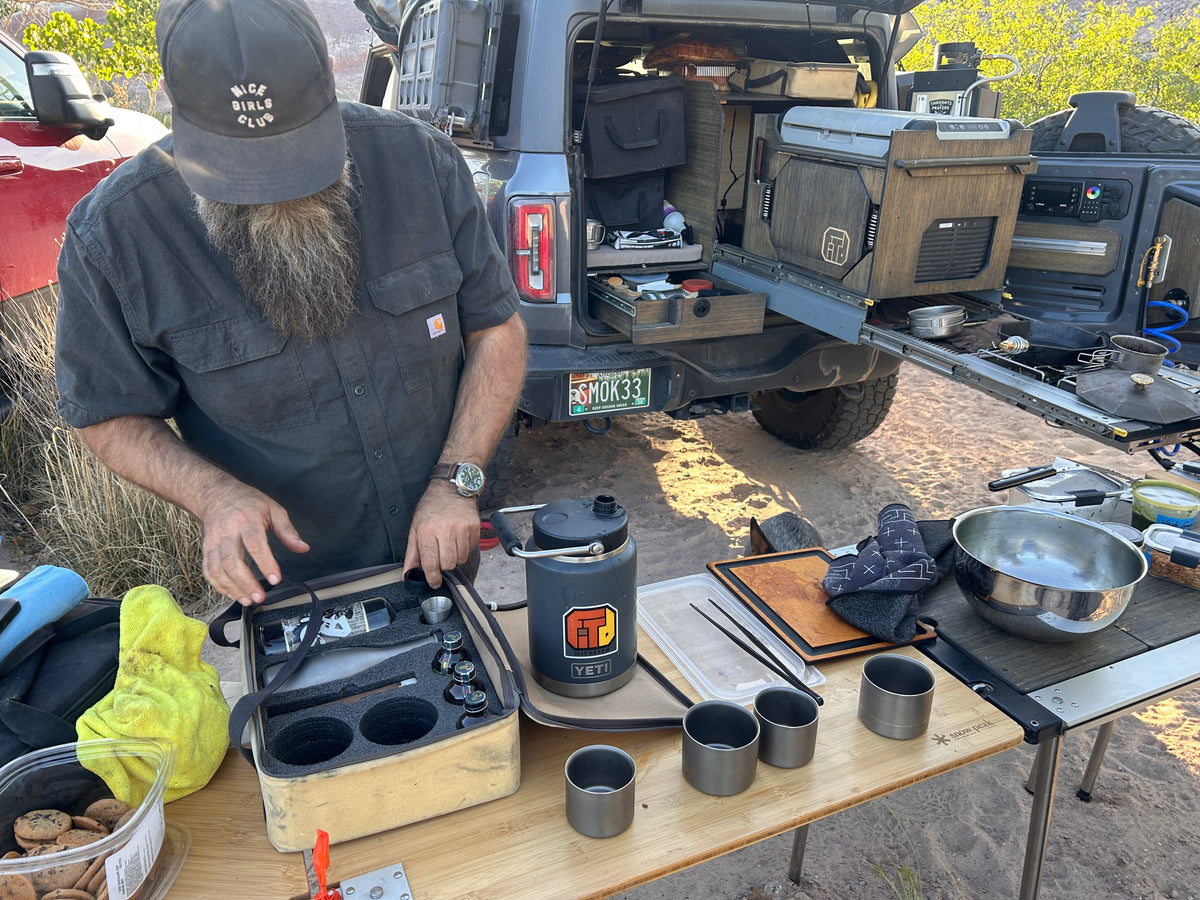 A man wearing all black stands over a long cooking table with a off-road equipped Bronco behind him. The man is setting up cooking utensils and pans to prepare a meal in a remote location.