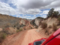 The view from the windshield of a red truck that is driving along a dusty dirt road winding through a rugged mountain landscape under a cloudy blue sky.