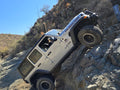 A silver Jeep Wrangler Rubicon drives up a very steep cliffside at an extreme angle with cactus and rocks in the background.
