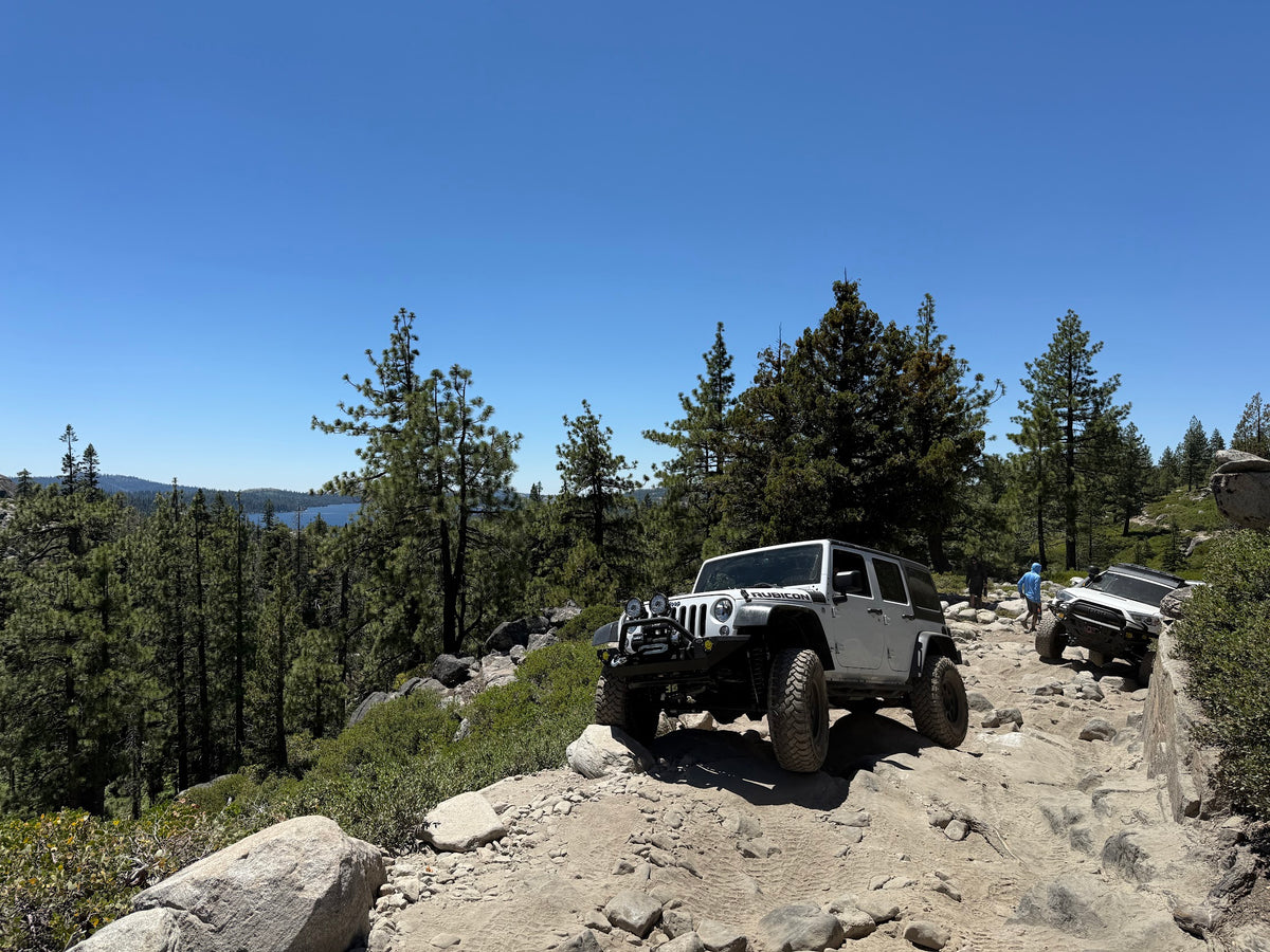 A white Jeep Wrangler is shown driving on a rocky mountain trail, with another vehicle behind it and a forested landscape under a clear blue sky.