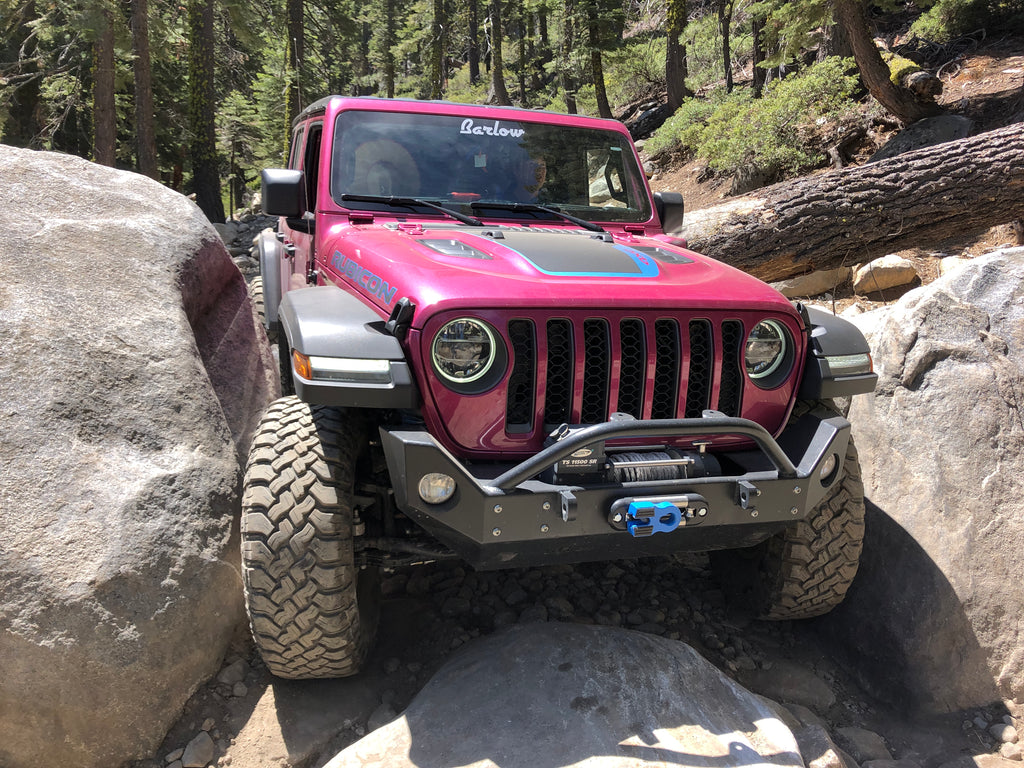 A pink Jeep Wrangler squeezes through a very tight opening between two large boulders with green trees in the background.