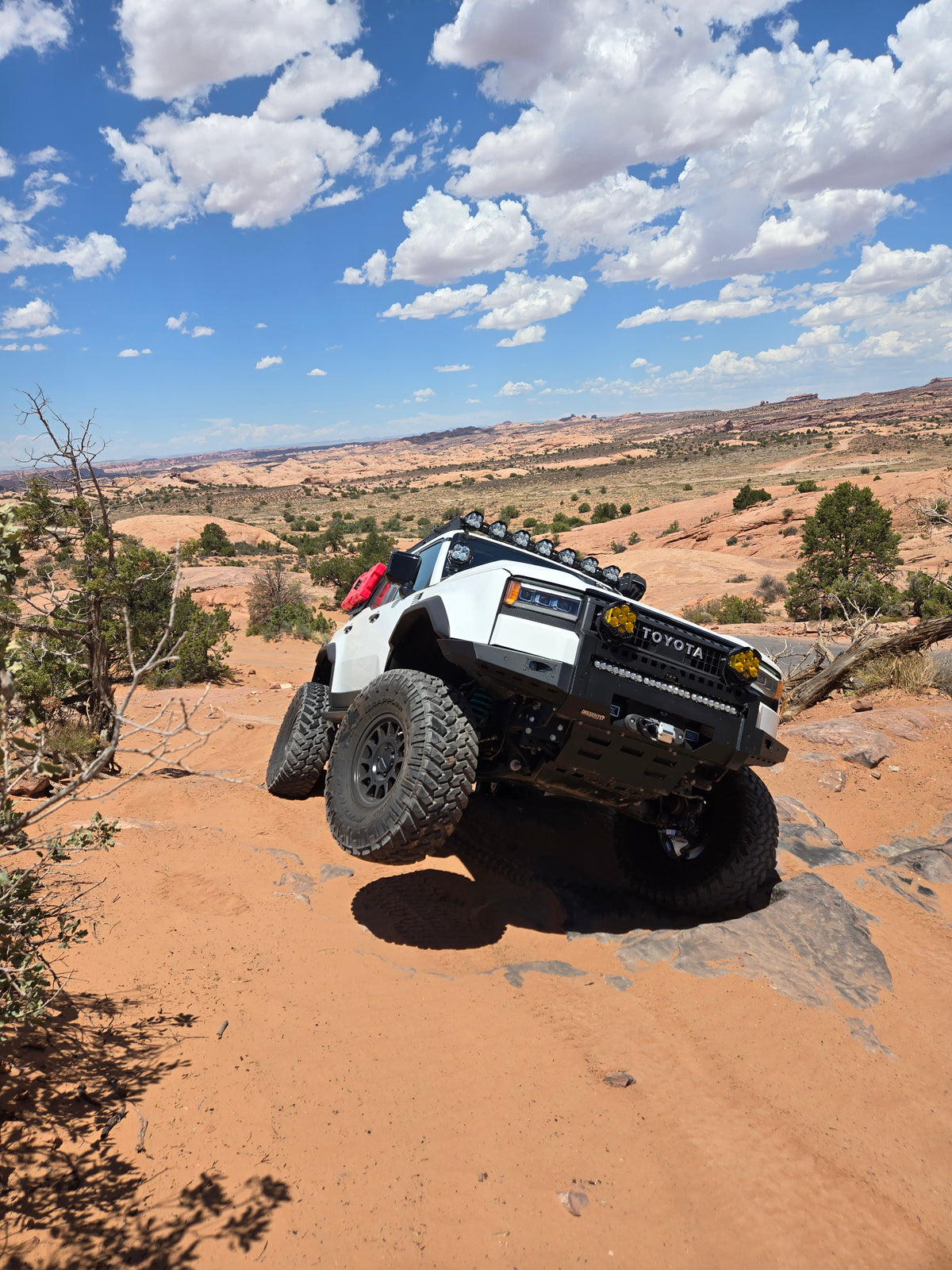 A white and black Toyota Land Cruiser  with large off-road tires is shown driving on a rocky desert trail under a blue sky with white clouds. The front passenger tire is raised in the air as the vehicle drives up a rocky and sandy slope.