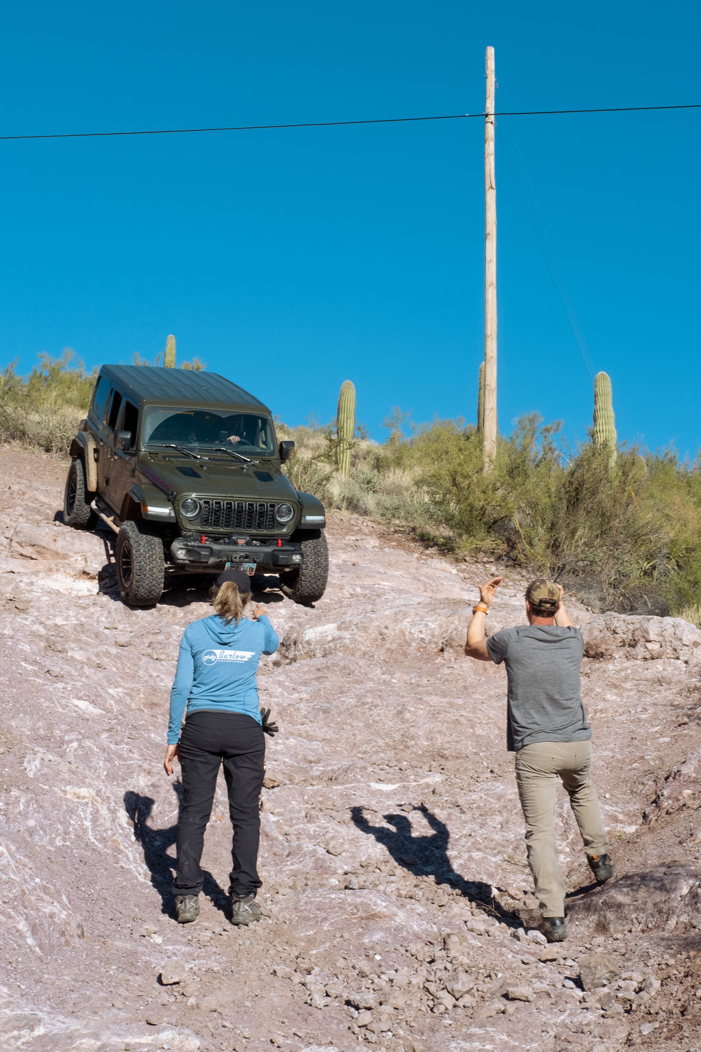 A Jeep Wrangler descends a steep dirt hill under a clear blue sky, with two people standing below on the slope giving hand signals. Saguaro cacti and shrubs are visible in the desert landscape.