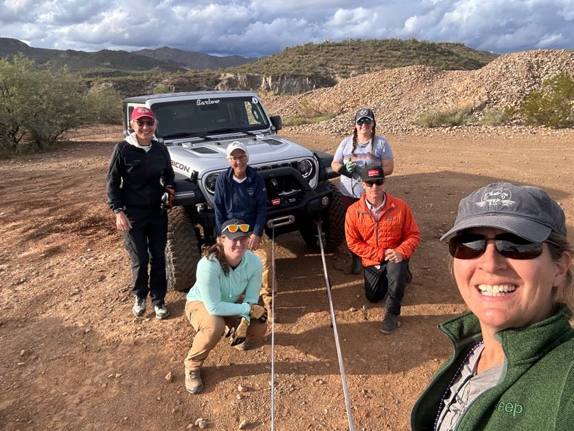 A group of six people pose outdoors in a desert setting with a silver off-road Jeep behind them. The group is dressed in casual outdoor apparel, and the background features rocky hills and a cloudy sky.