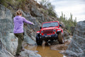 A red off Jeep is navigating a muddy desert wash in between large boulders as a woman in a purple hoodie and olive pants spots the driver through the tight area.
